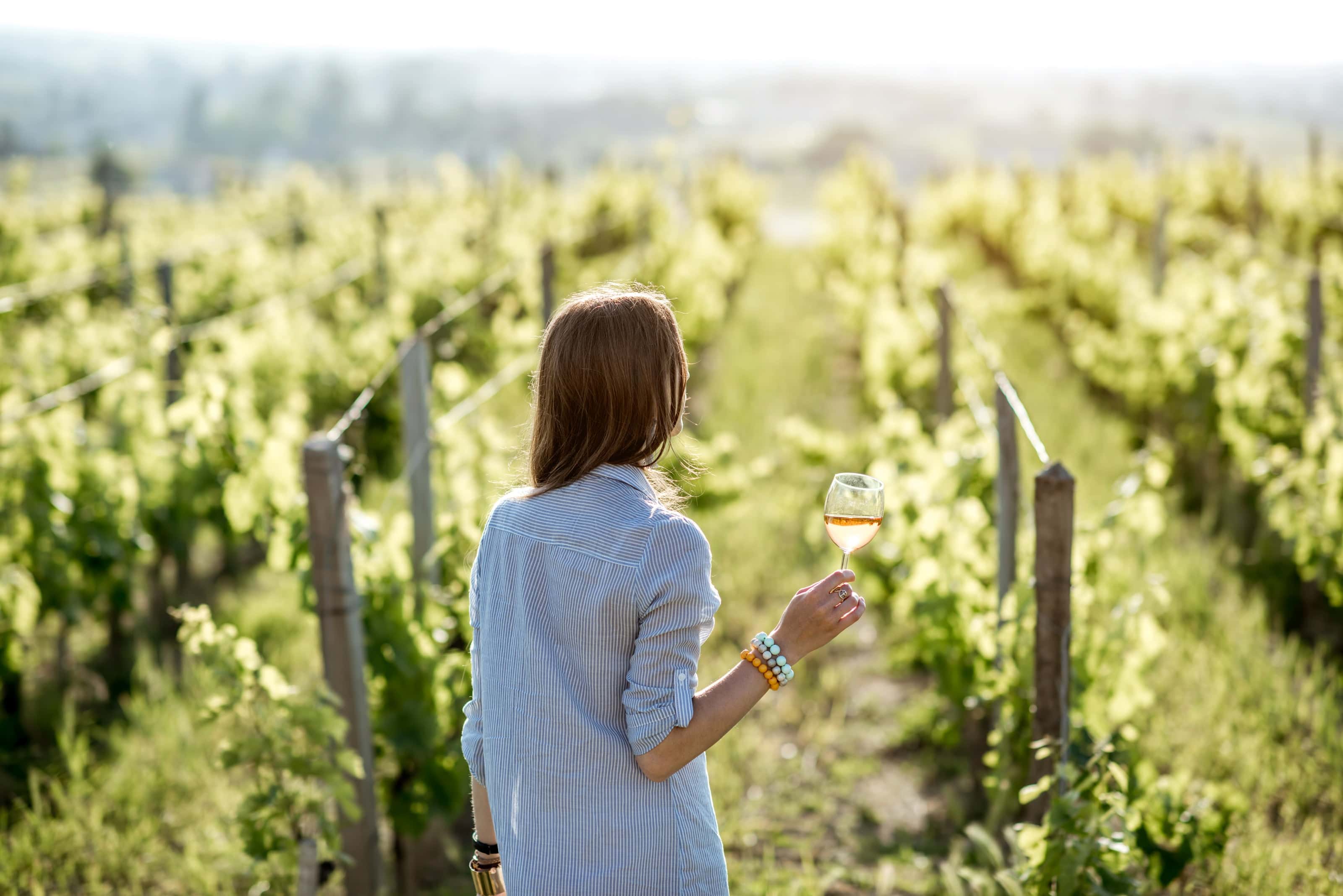 Young woman holding a glass of white wine while walking through a sunlit vineyard, symbolizing sustainability and women in winemaking.
