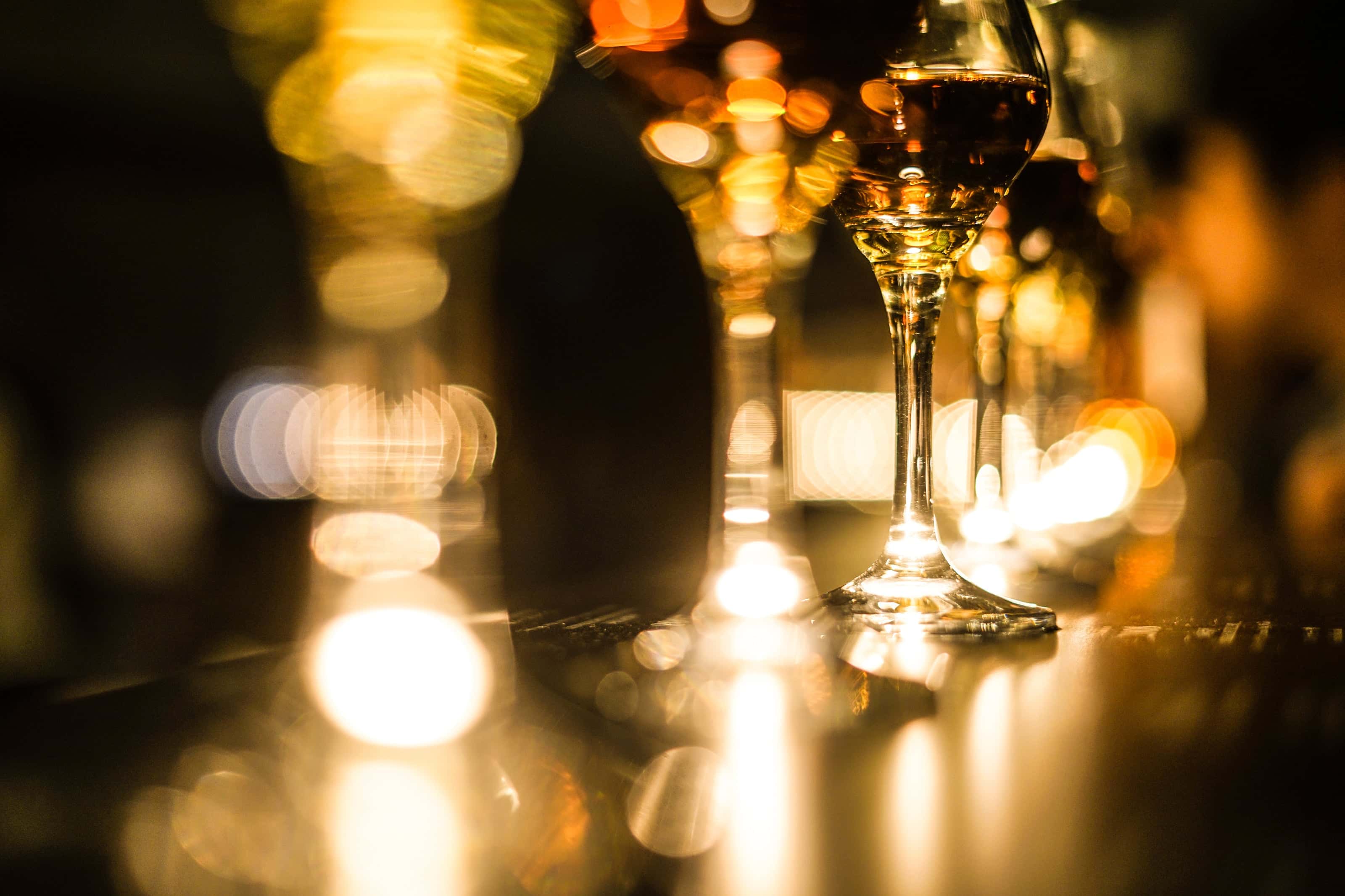 Close-up of wine glasses on a dimly lit bar counter with warm golden bokeh lights creating a festive ambiance.