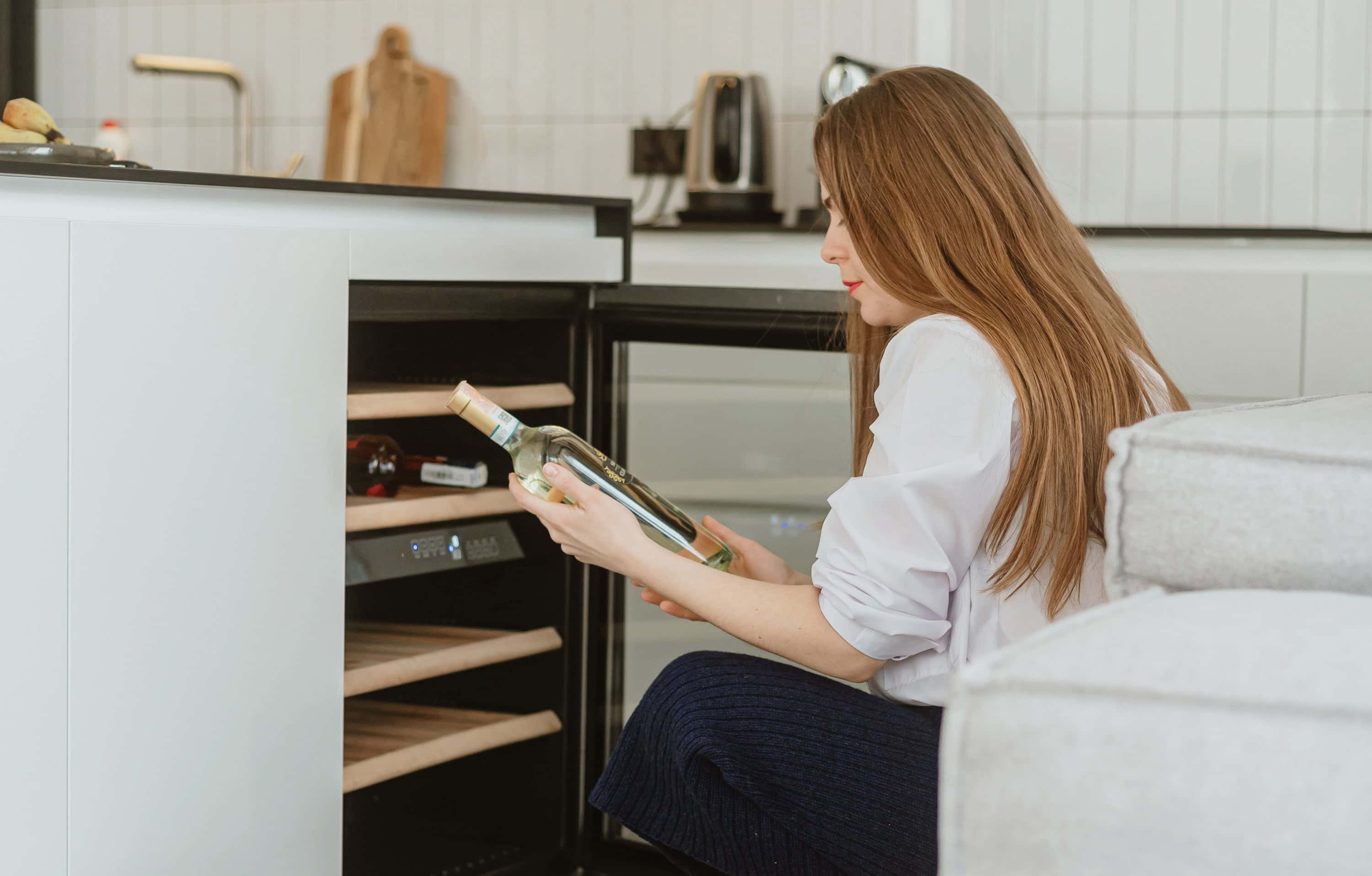 Woman kneeling in a modern kitchen while placing a bottle of white wine into a built-in wine fridge with wooden shelves.