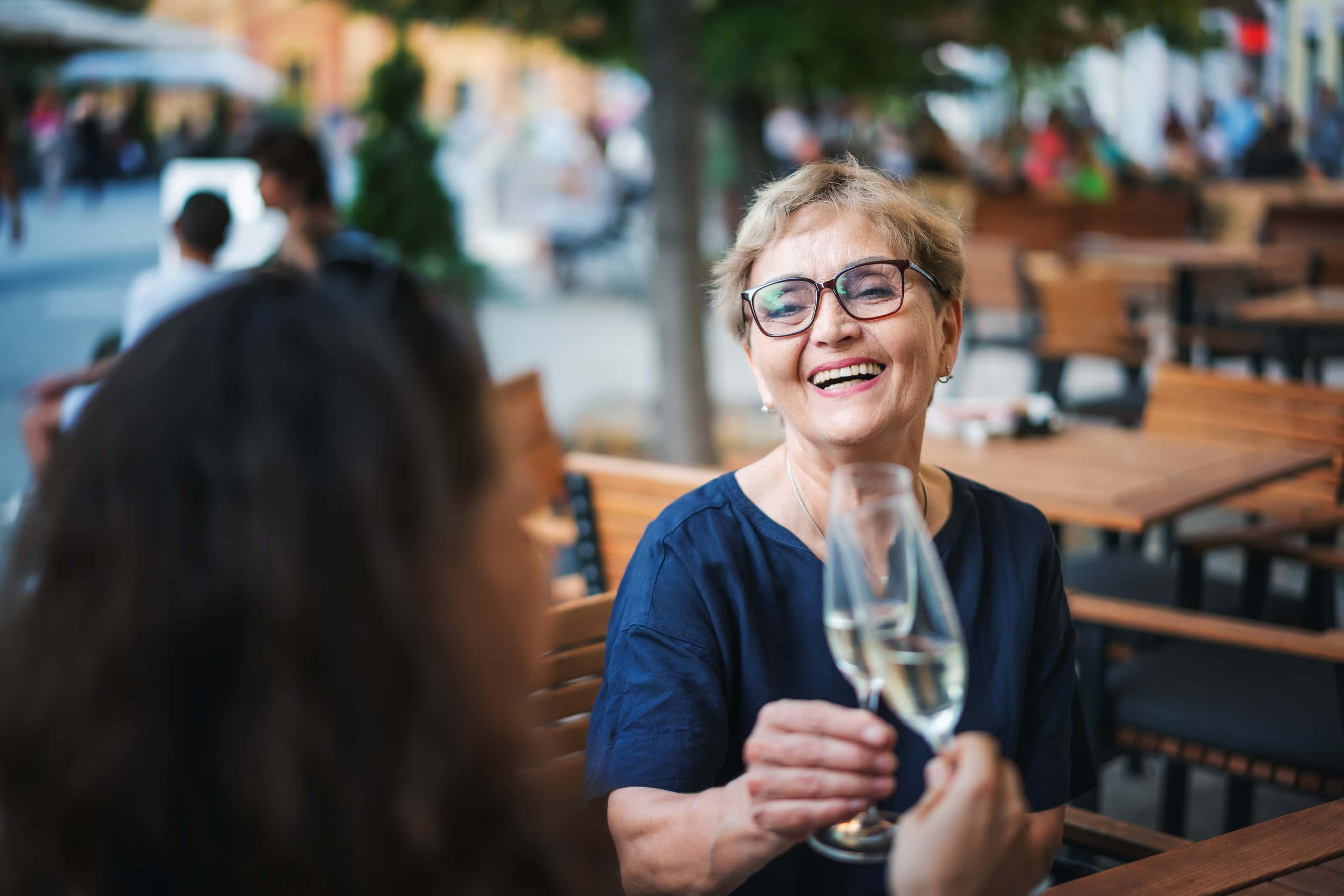 Smiling older woman clinking champagne glasses with a friend at an outdoor café, enjoying a lively social moment on a sunny day.