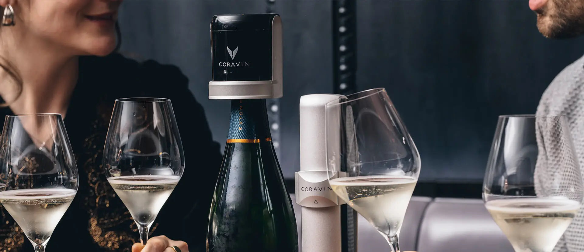 Two people enjoying glasses of sparkling wine at a table with a Coravin Sparkling Wine Preservation System attached to an open champagne bottle, showcasing a sleek and modern way to keep bubbles fresh.