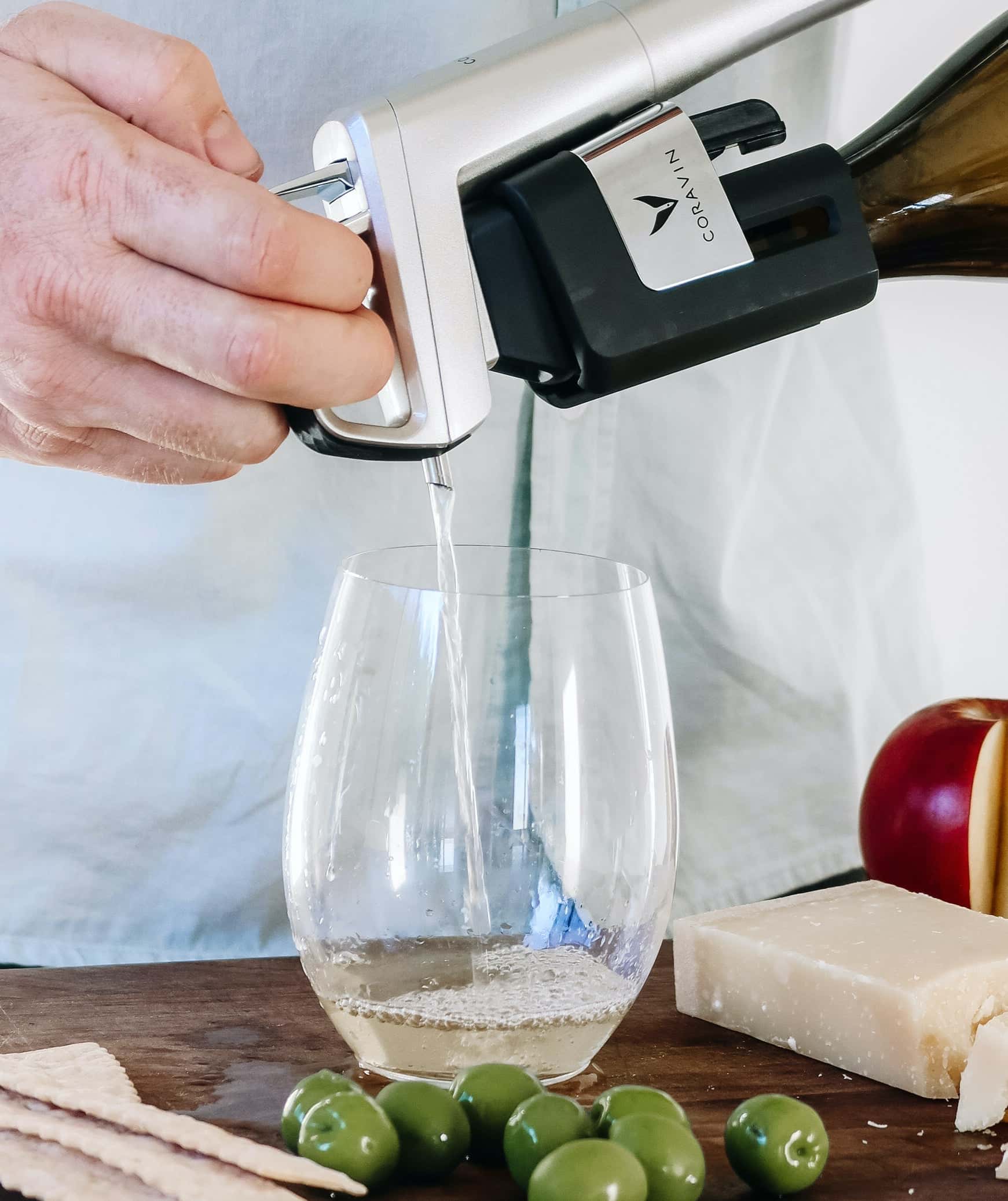 Close-up of a Coravin wine preservation system pouring white wine into a stemless glass, with green olives, crackers, Parmesan cheese, and sliced apple on a wooden surface.