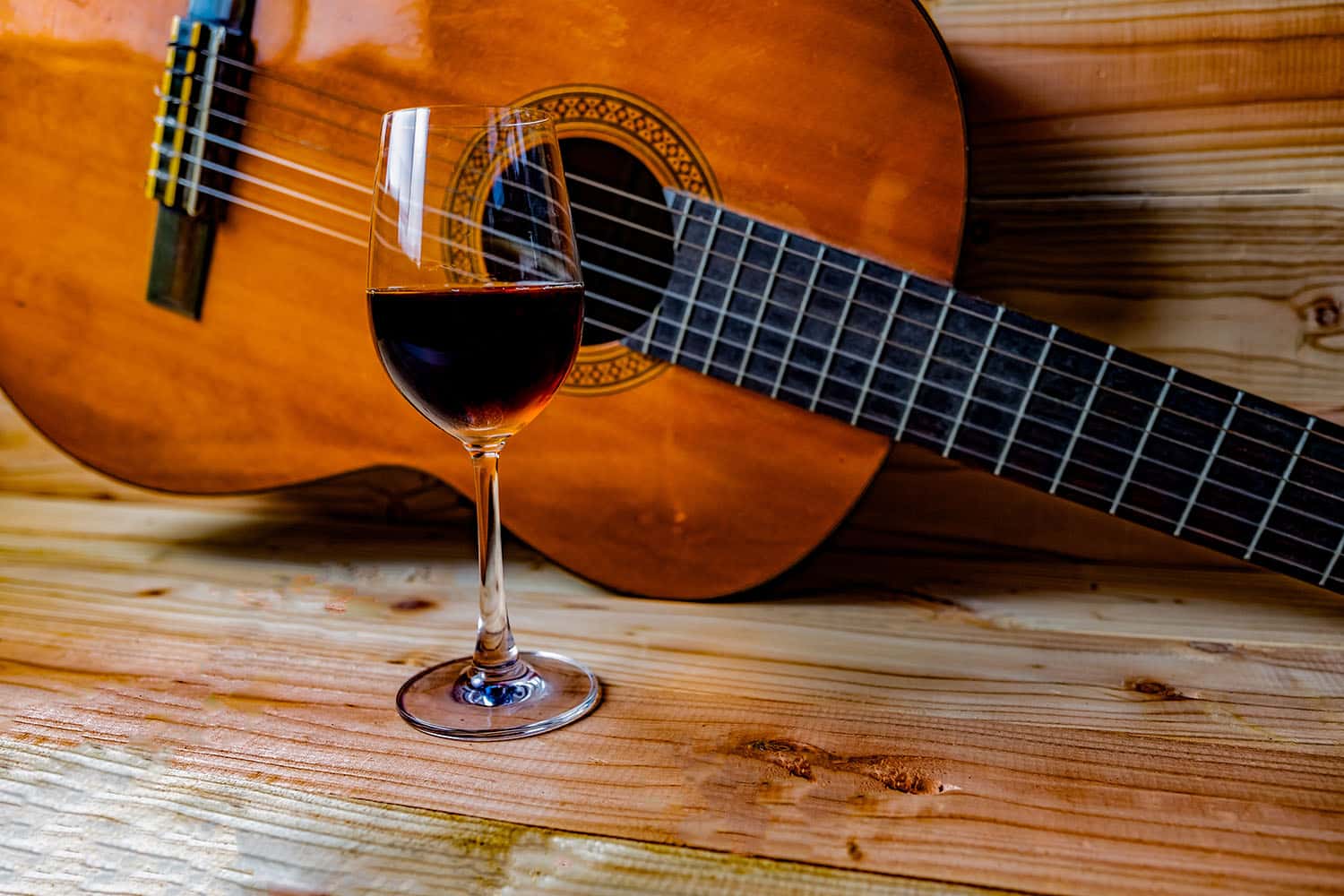 Glass of red wine placed on a wooden table next to a classical guitar, evoking a rustic and musical Spanish ambiance.