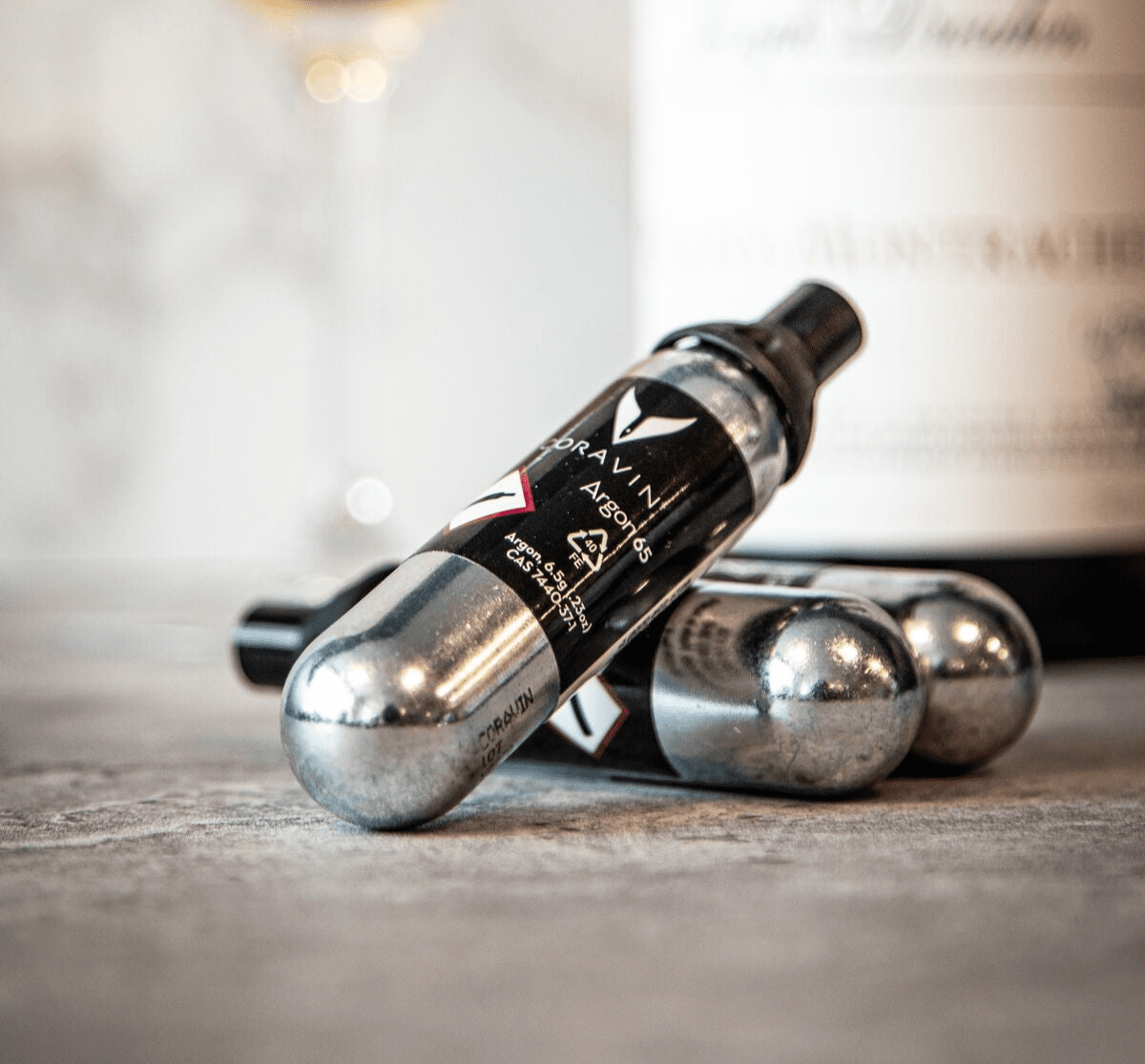 Three silver gas with black labels are arranged on a textured surface, with a blurred wine bottle in the background.