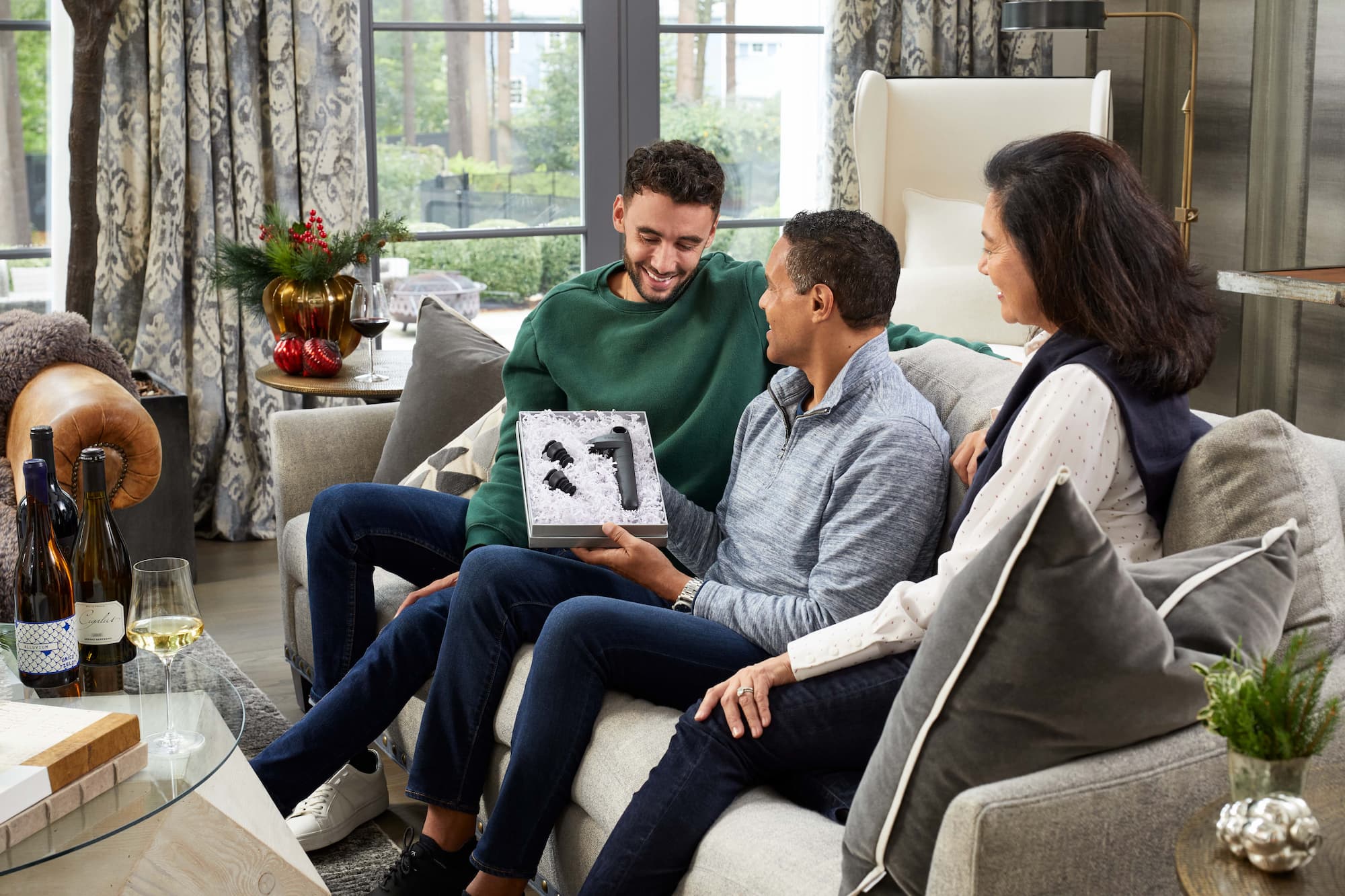 Three people exchanging gifts in a cozy, upscale living room decorated. One person holds a gift box with a coravin wine preservation system