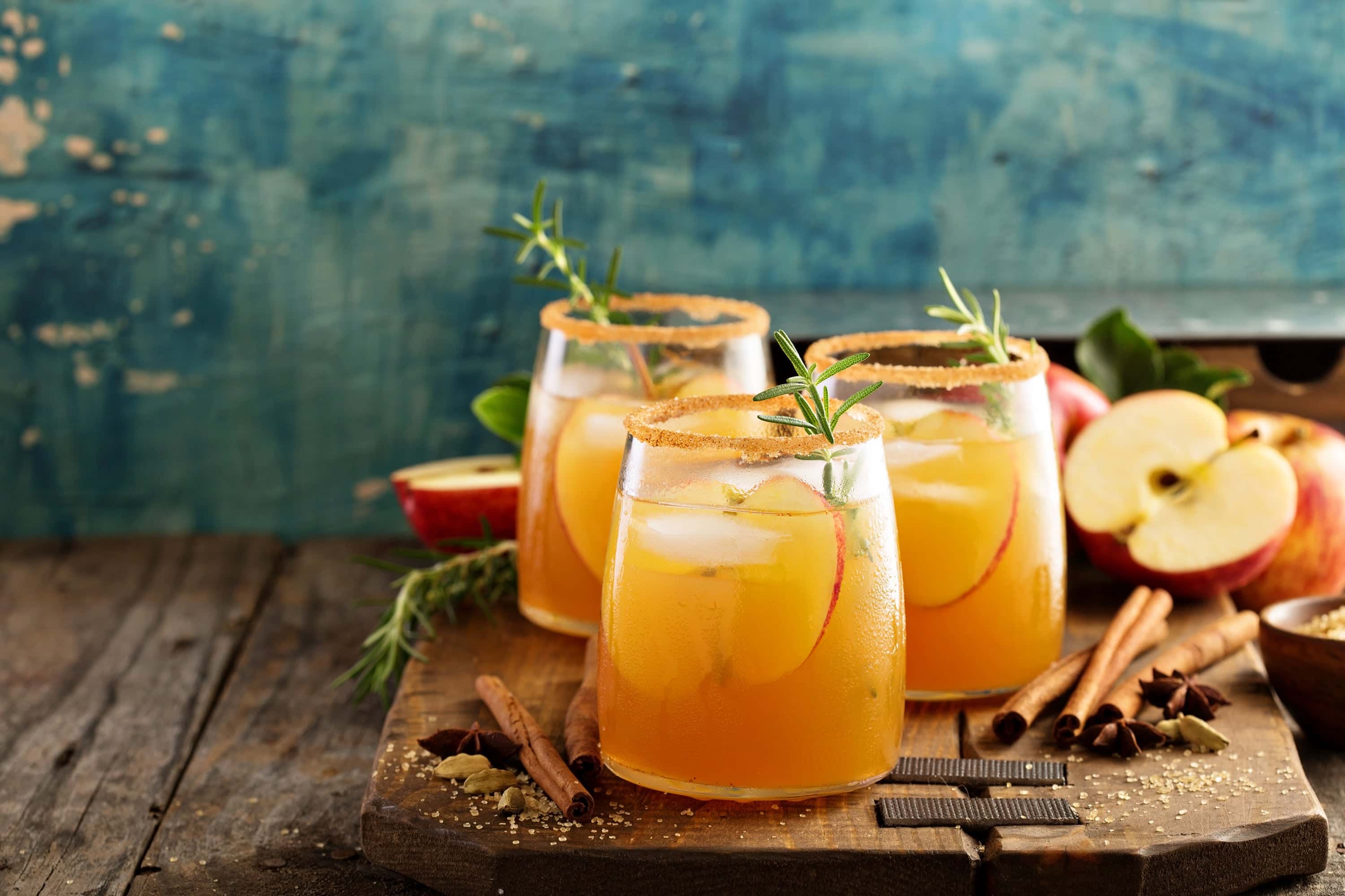 Fall apple cider cocktails with cinnamon sugar rims, garnished with apple slices and rosemary, served on rustic wooden board with spices and fresh apples.