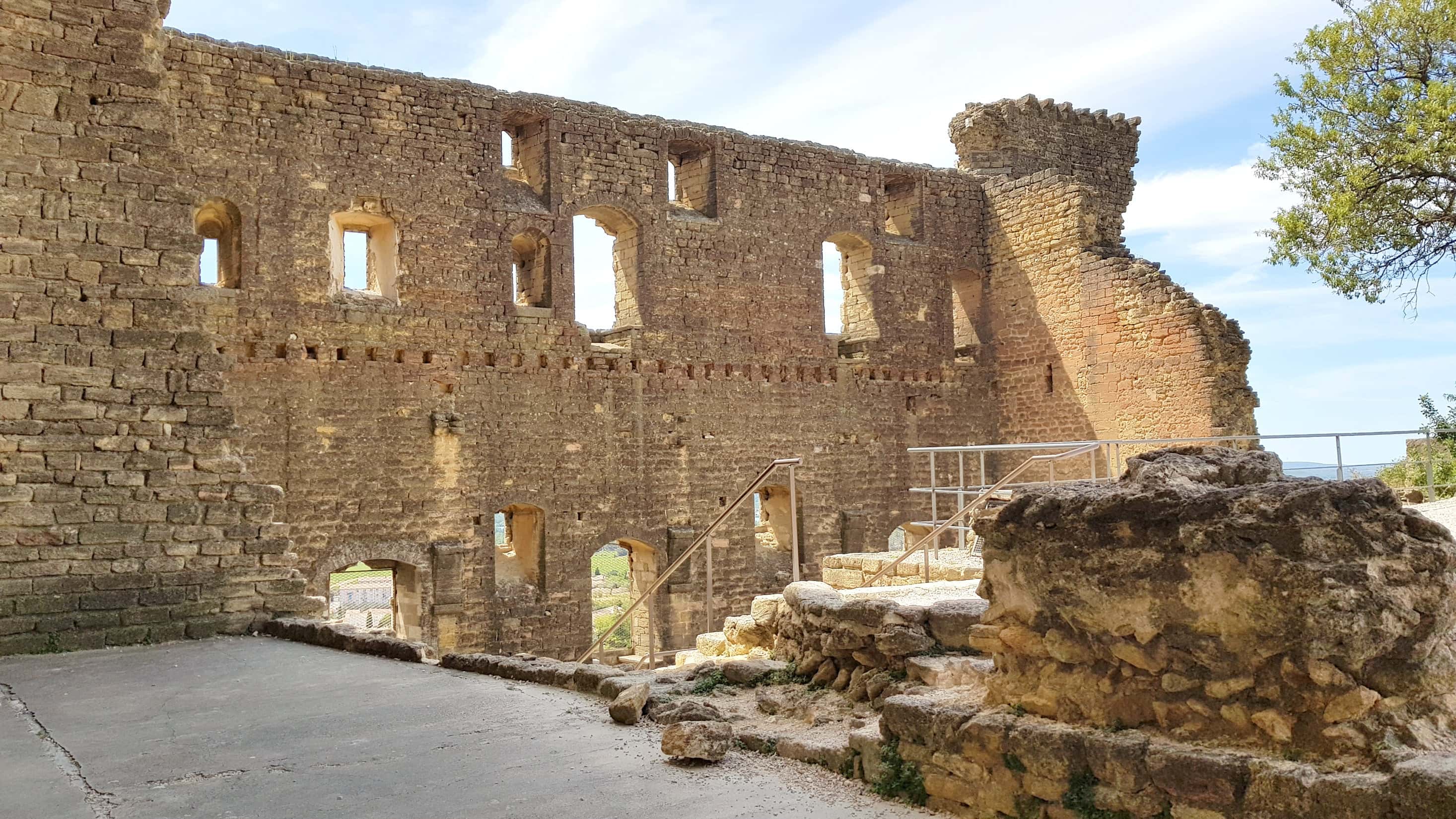 Ruins of a medieval stone castle with arched windows and a crumbling tower under a blue sky, located in a historic European countryside.