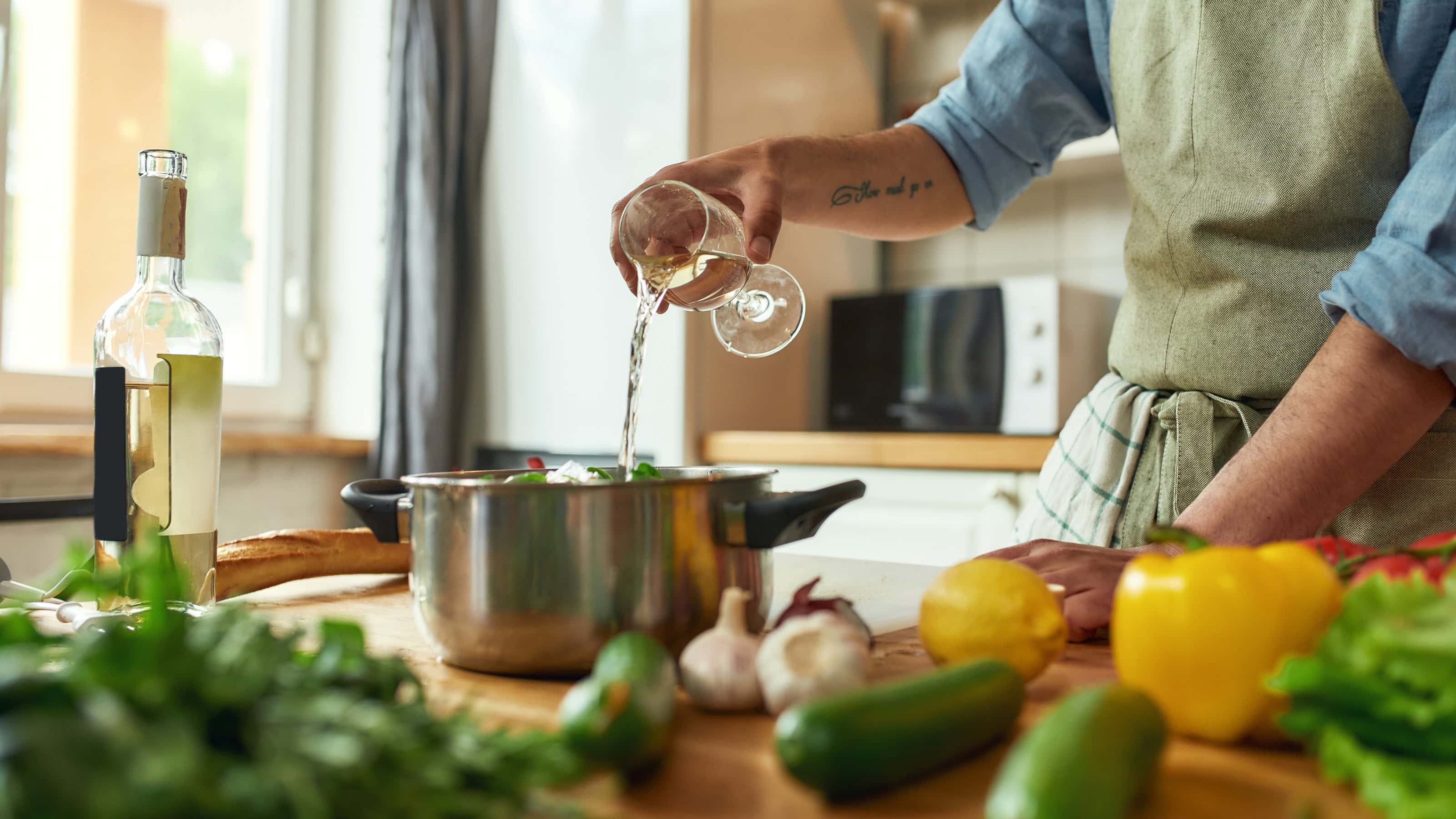 Home cook pouring white wine into a pot surrounded by fresh vegetables, showcasing Coravin for cooking with wine in a bright kitchen.