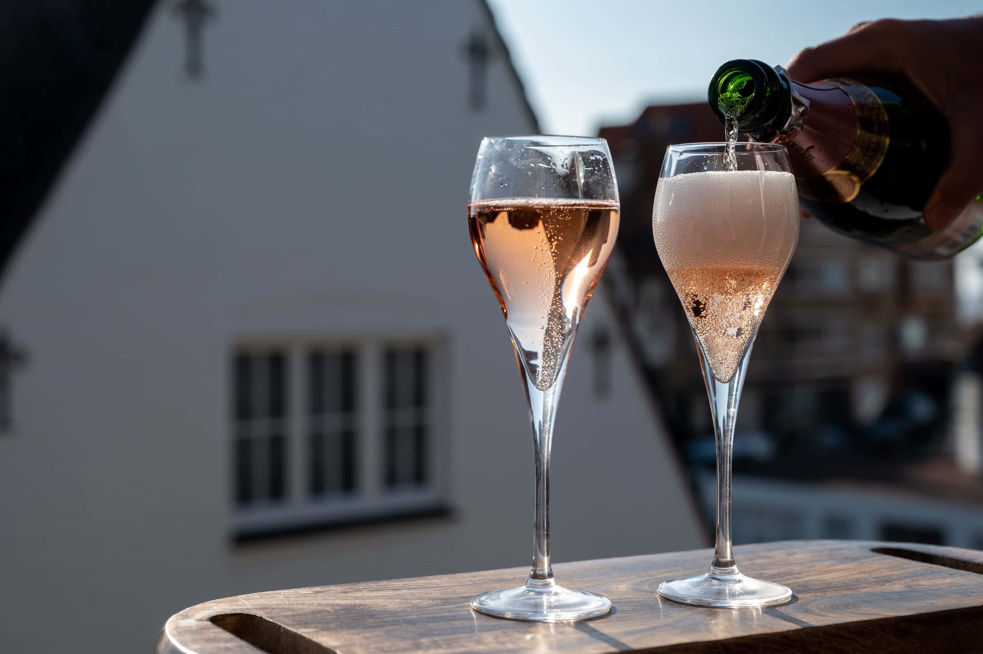 Cava wine being poured into elegant champagne flutes on an outdoor table, with a soft-focus background of urban rooftops and evening light.