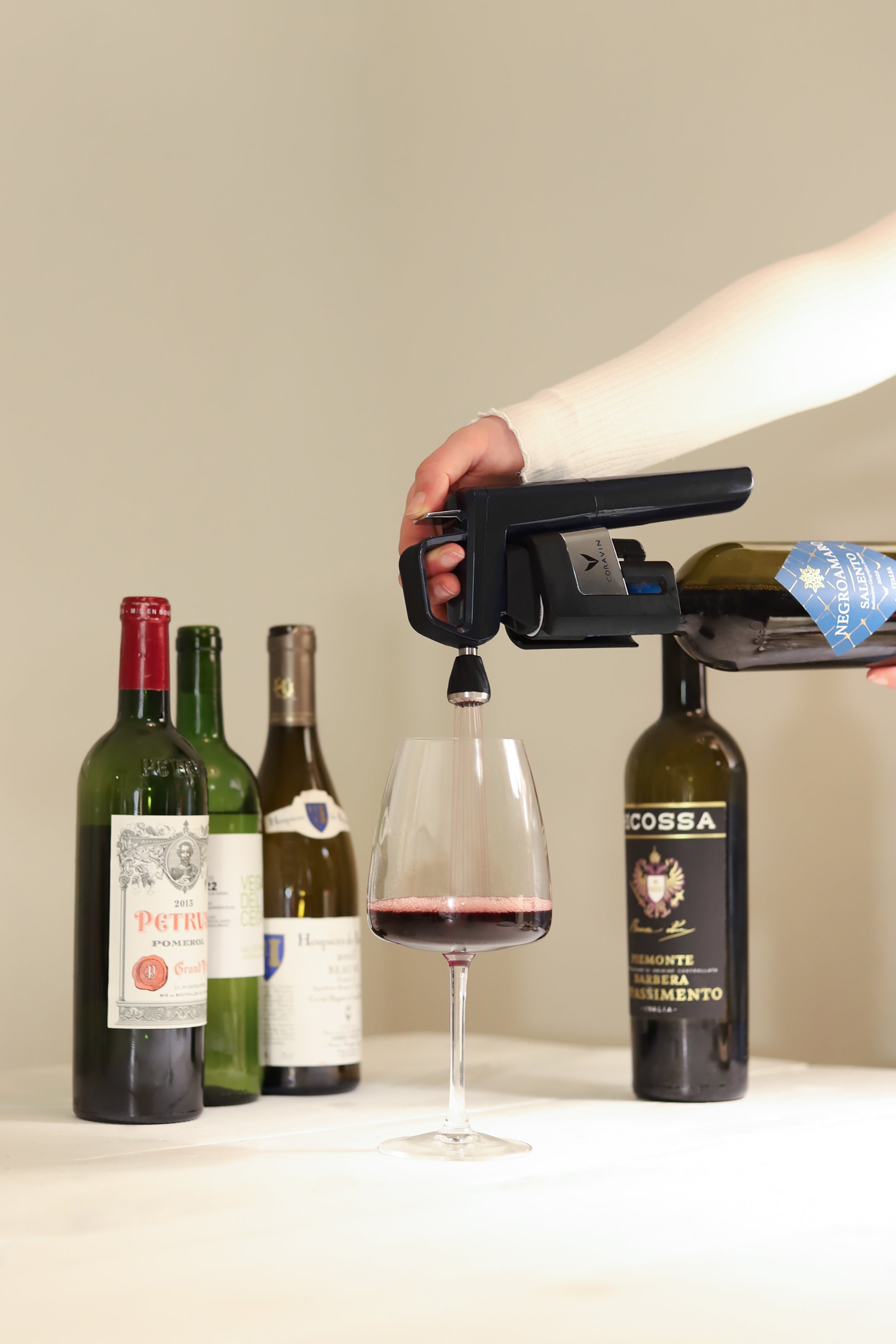 Person using a Coravin Timeless wine preservation system to pour red wine from a bottle into a glass, with several premium wine bottles displayed in the background.