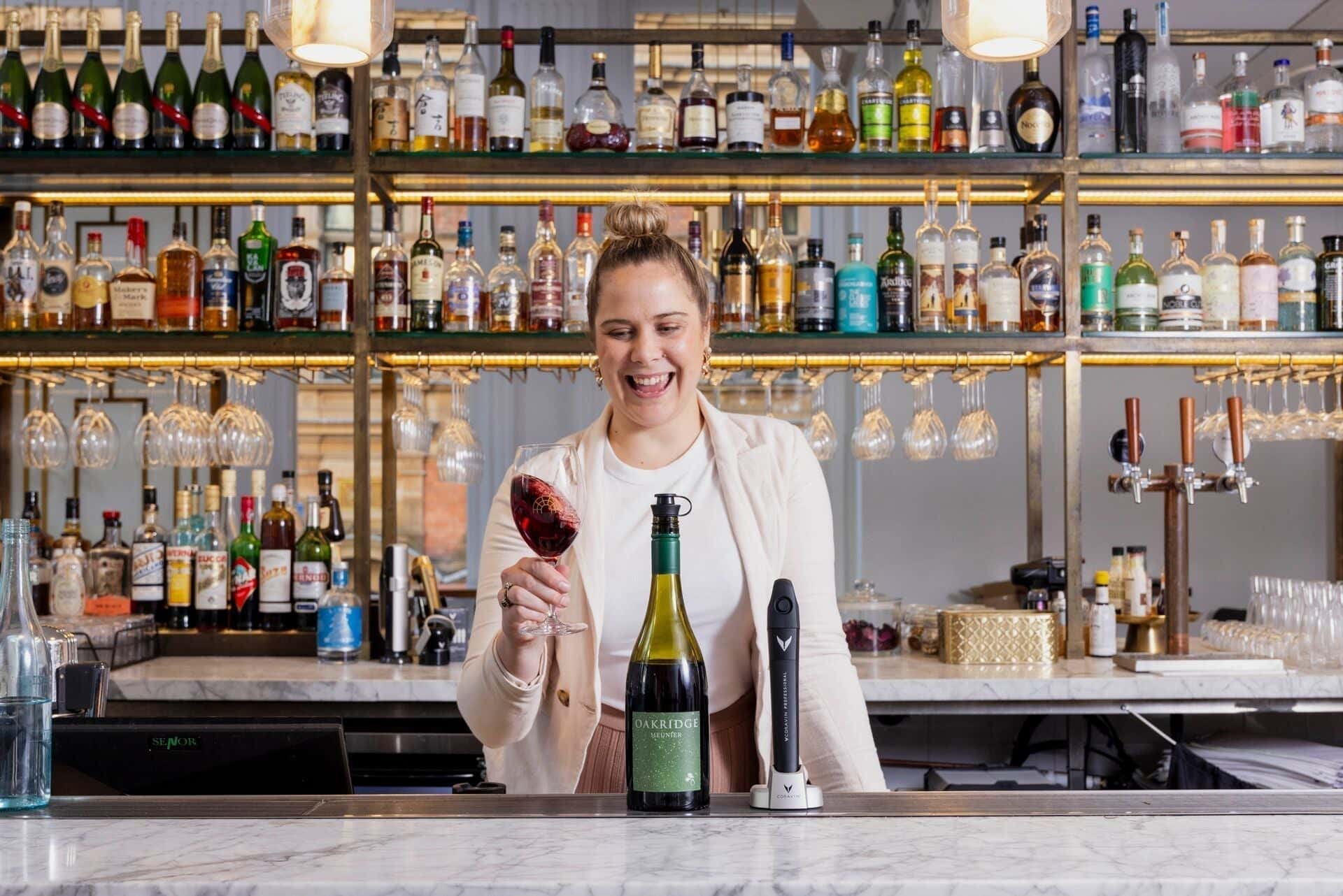 Sommelier Louella Mathews using a Coravin wine preservation system behind a bar, holding a glass of red wine with a bottle of Oakridge Pinot Noir.