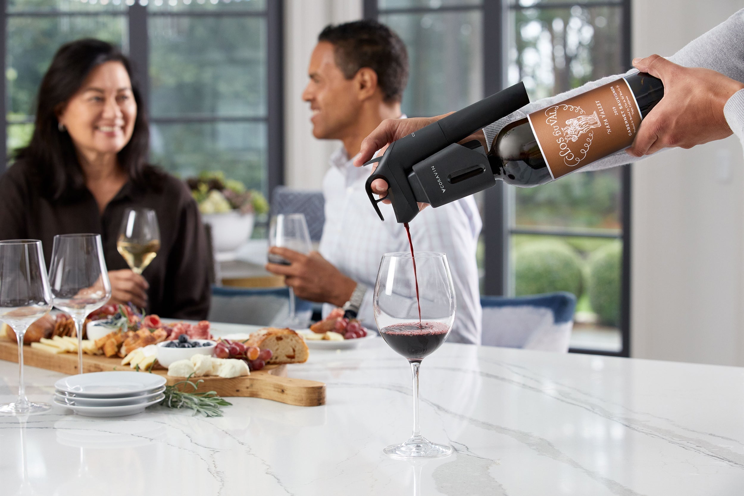 Person using Coravin wine preservation system to pour red wine while friends enjoy a charcuterie board and white wine at a modern kitchen gathering