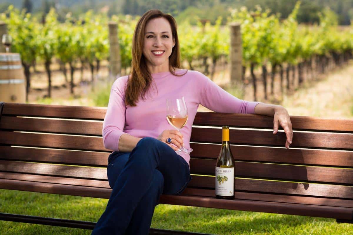 Woman relaxing on a bench in a vineyard, holding a glass of white wine with a wine bottle beside her.