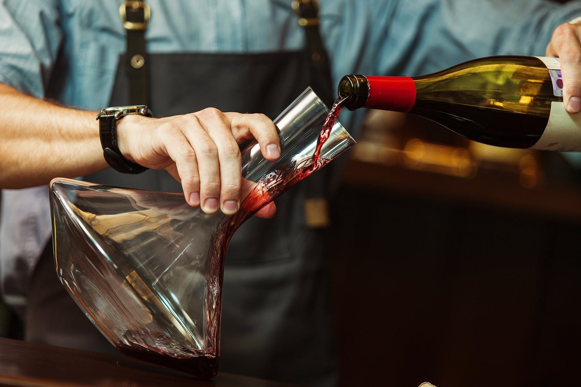 Sommelier slowly pouring red wine from a bottle into a modern angled wine decanter to aerate and separate sediment.
