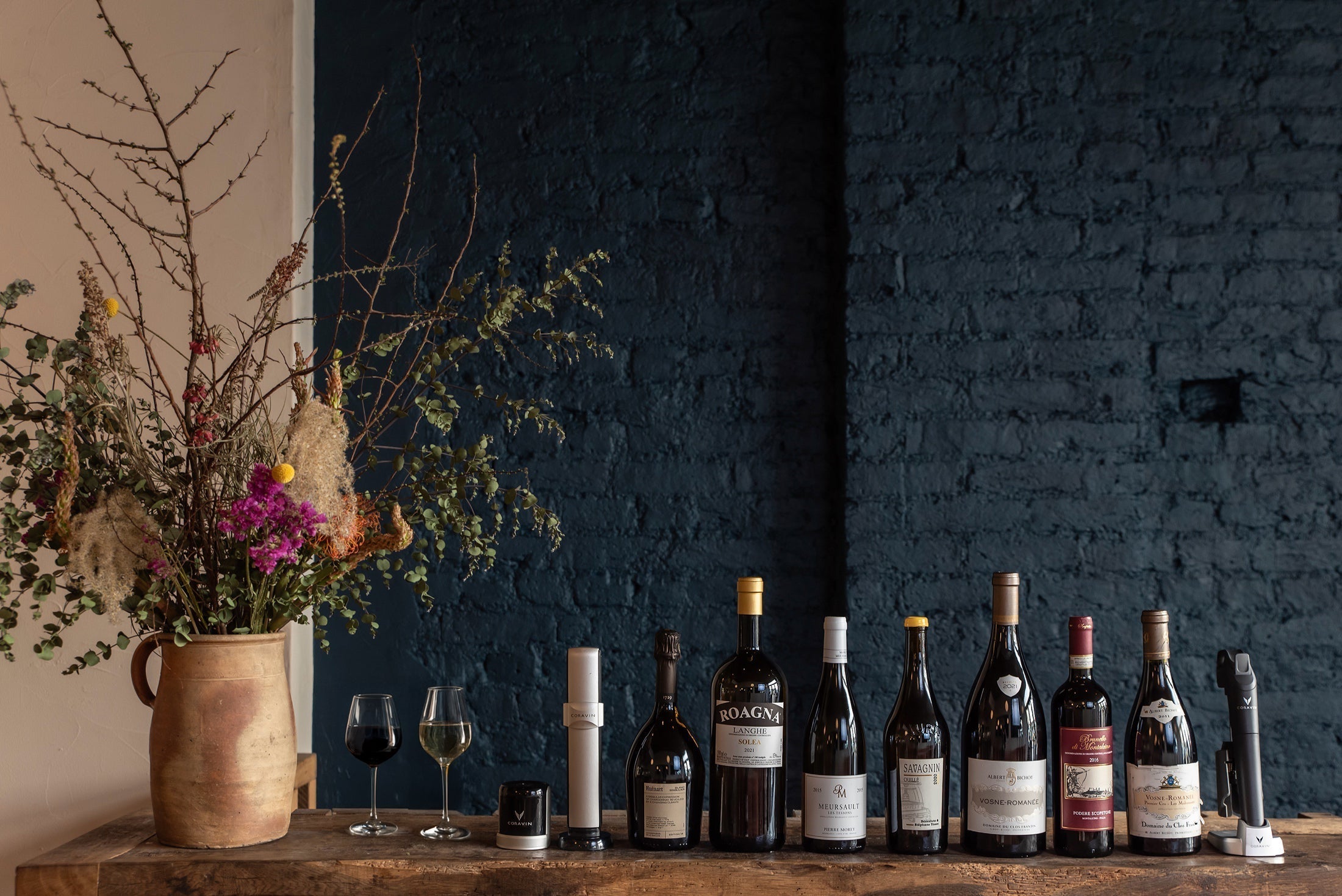Selection of fine wines with Coravin wine preservation systems on rustic wooden table beside floral arrangement, set against a dark blue brick wall