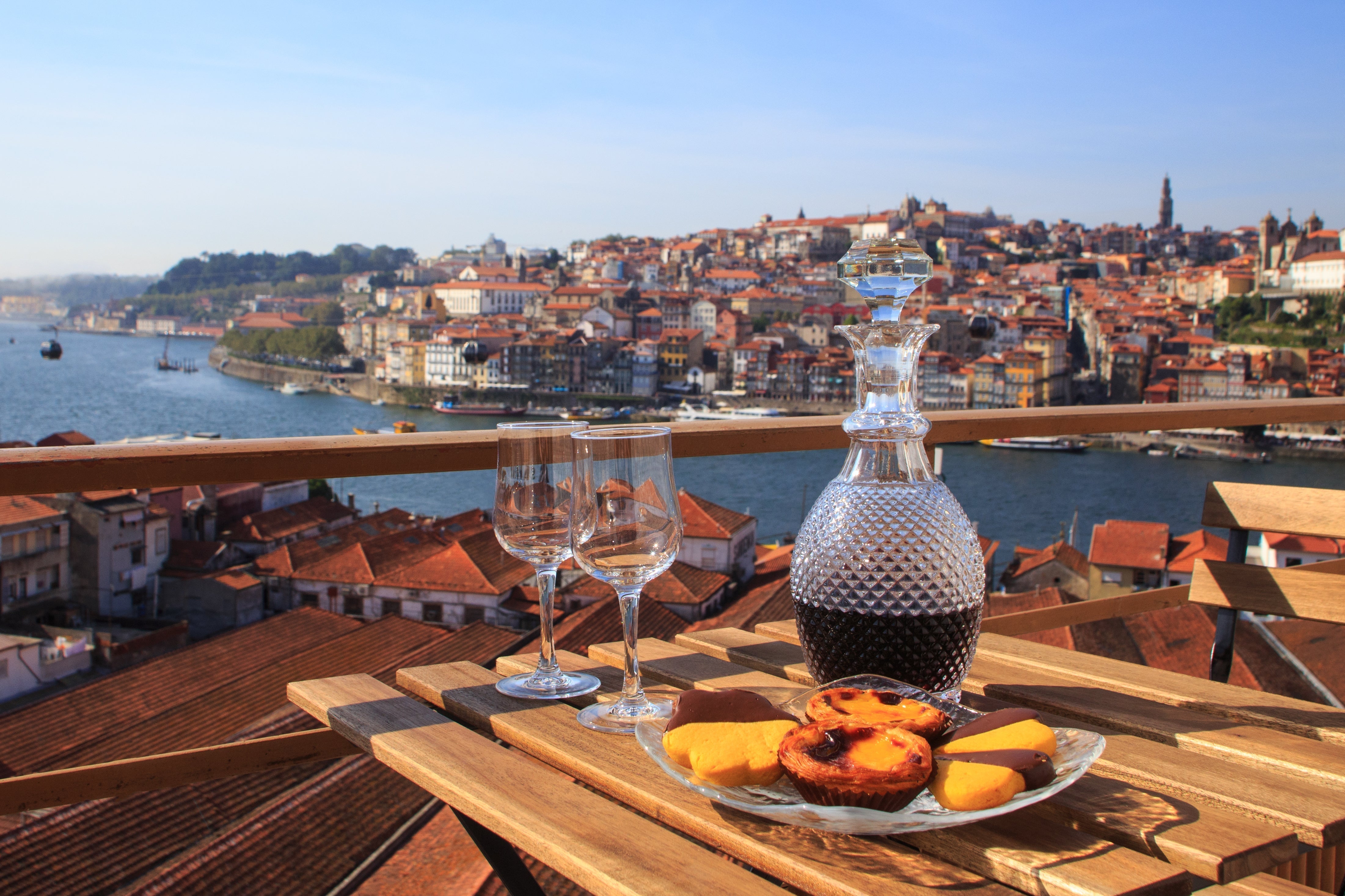 Port wine and traditional Portuguese pastries on a wooden table overlooking the Douro River and the historic cityscape of Porto, Portugal.