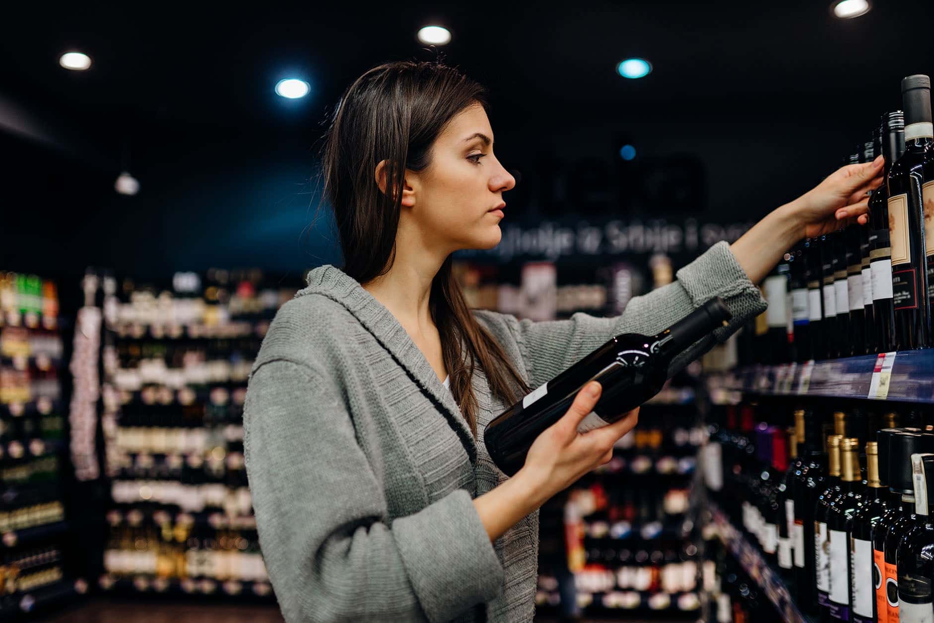 Young woman selecting a bottle of wine from a shelf in a liquor store, carefully examining wine labels under bright store lighting.