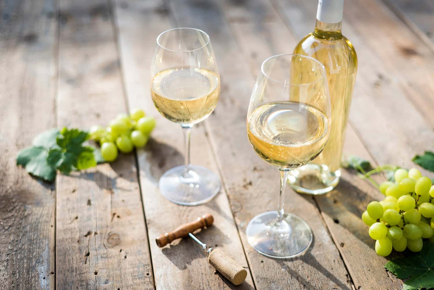 Two glasses and a bottle of chilled white wine on a rustic wooden table, surrounded by green grapes, grape leaves, and a corkscrew—perfect for an outdoor wine moment.