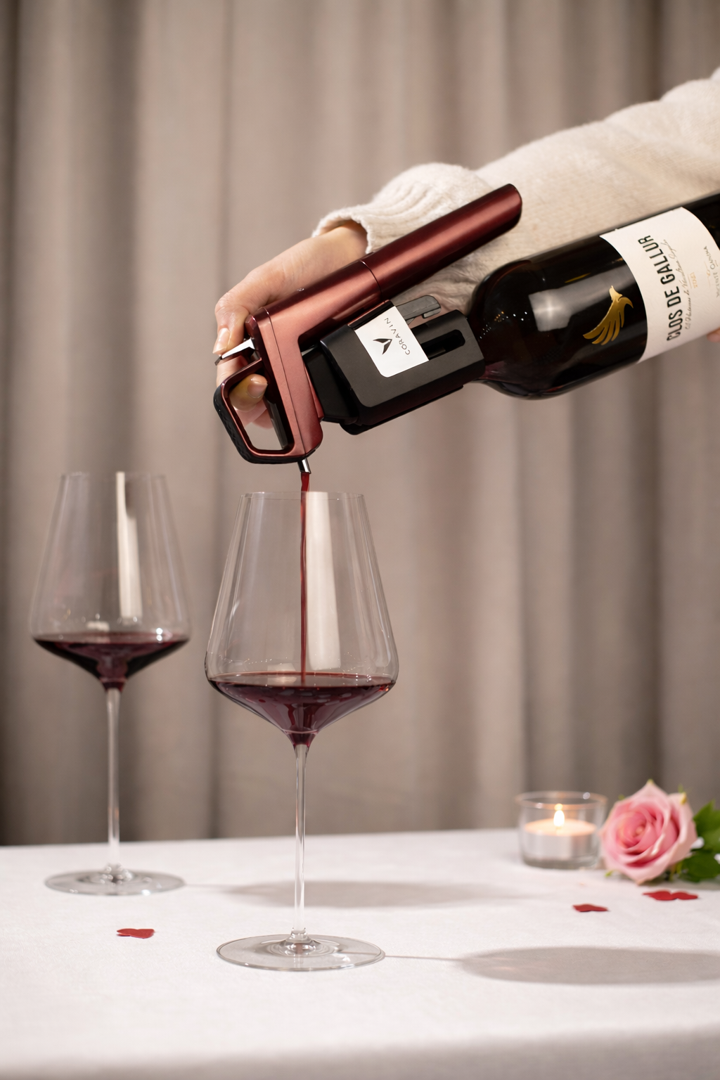 Person pouring red wine from a bottle into a glass on a table with a candle and flower.