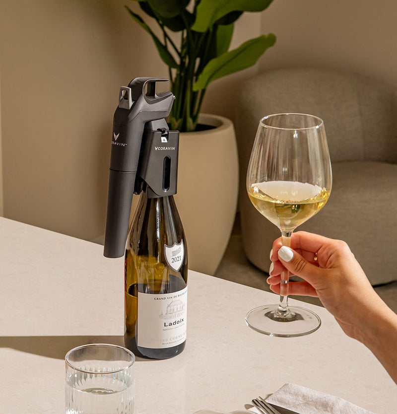 Person holding a glass of white wine next to a bottle and wine opener on a table.