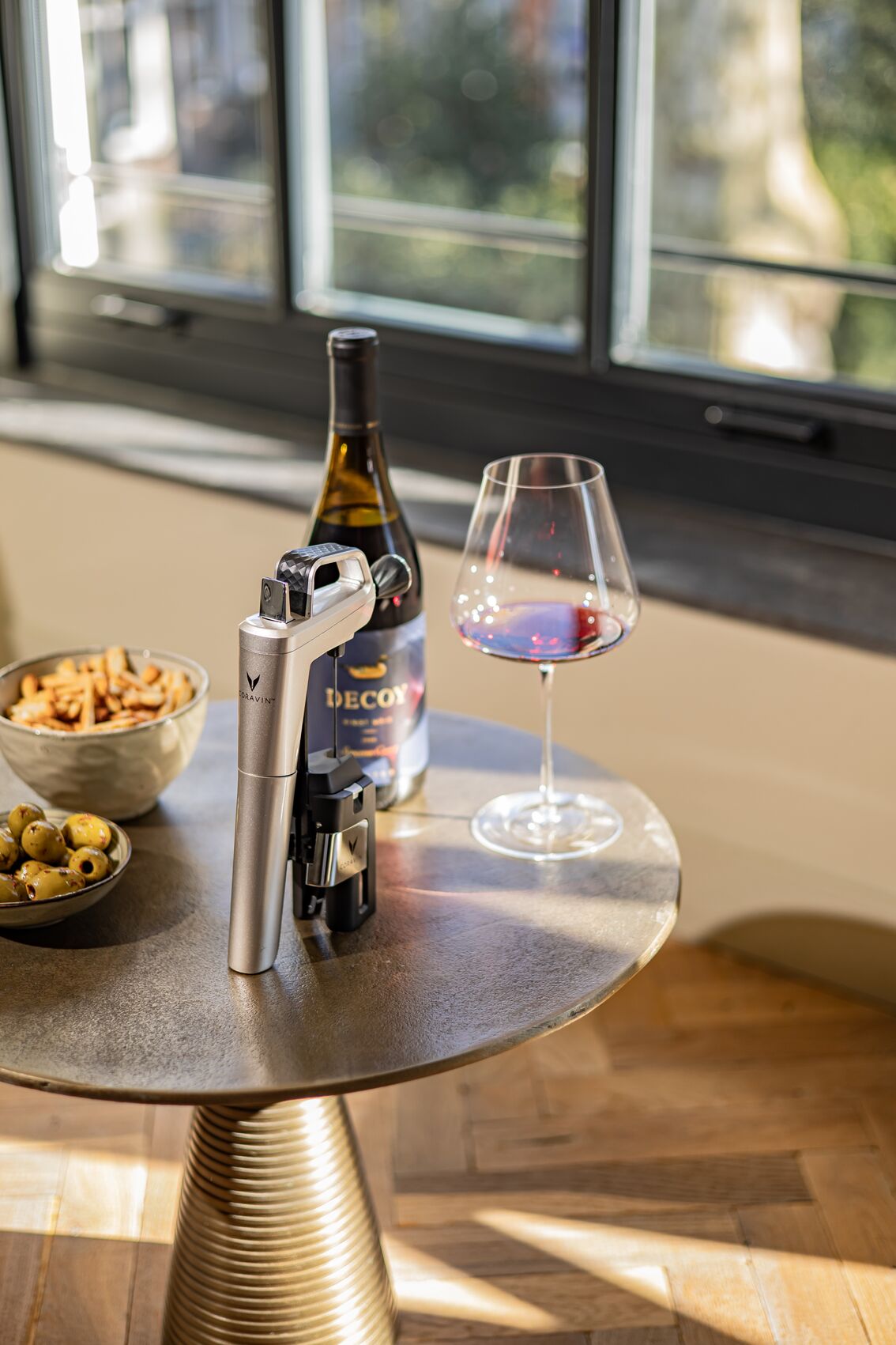 Wine bottle, glass, and Coravin Timeless Six in Silver on a table with snacks in a living room with windows.