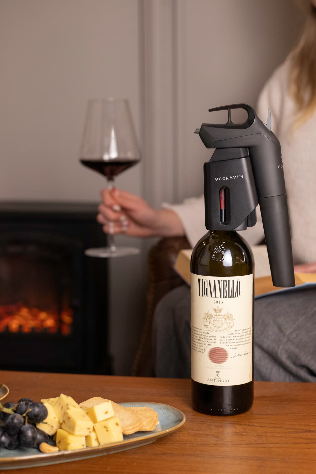 A bottle of 2019 Tignanello red wine with a Coravin preservation system attached sits on a table with a plate of assorted cheeses, crackers, and grapes