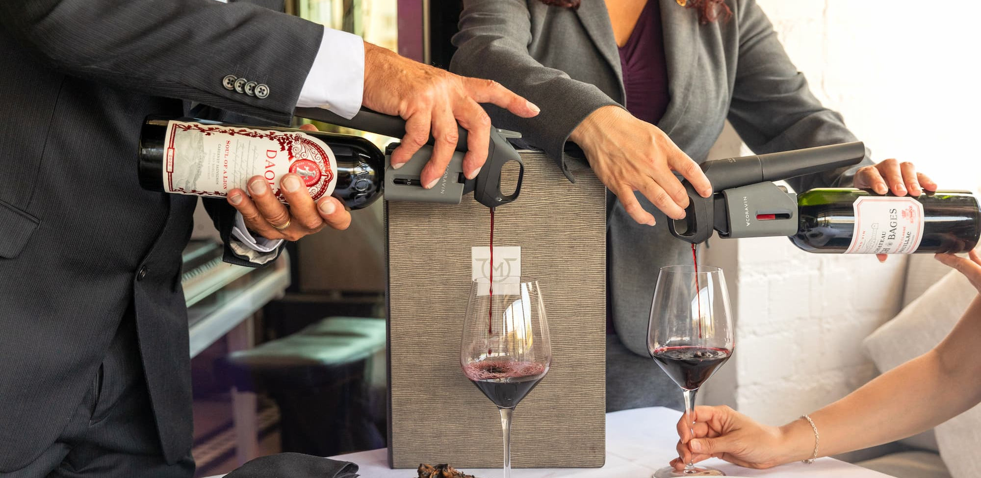 a small party of people enjoying a fine dining experience, with two using Coravin wine preservation systems to pour red wine into glasses without removing the cork