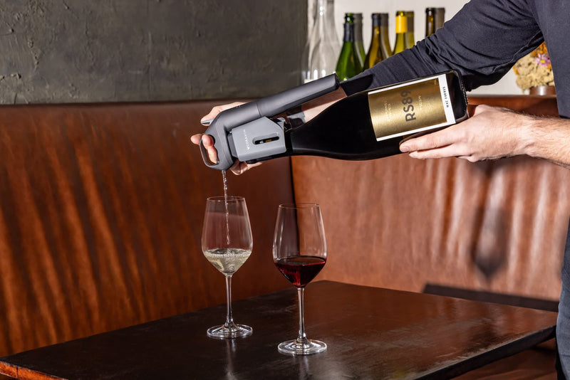 A person using a Coravin wine system to pour white wine from a sealed bottle with another glass of red wine, evoking a refined, modern wine-tasting setting